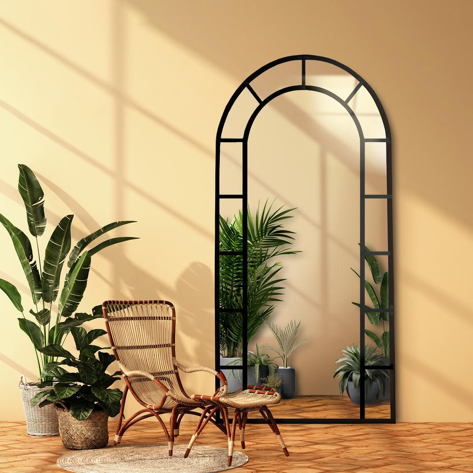 Ein großes, bogenförmiges Spiegel mit schwarzem Metallrahmen, das an eine beige Wand gelehnt ist, umgeben von grünen Pflanzen und einem Rattanstuhl im Boho-Stil, mit warmem natürlichem Licht.